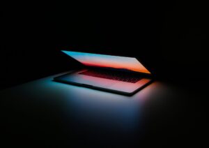 Nell'immagine si vede un computer portatile quasi del tutto chiuso. Lo schermo è acceso e illumina scarsamente la tastiera con una luce blu e rossa sfumata. Tutto intorno è buio.
