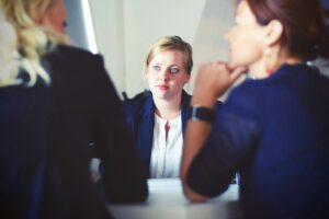 immagine rappresentante 3 business women: punto di vista tra due donne di schiena (da dietro sopra le loro spalle) con in mezzo la terza, vista di fronte, al dia disuna scrivania che le separa.