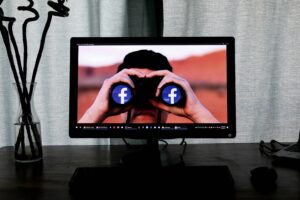Immagine di un monitor di un pc fisso su una scrivania, con uno screensaver che rappresenta una persona con un cannocchiale le cui lenti rappresentano il logo di Facebook.