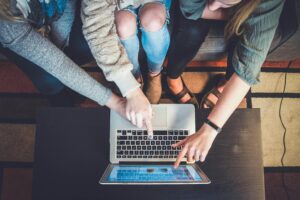 persone viste dall'alto, sedute su un divano, che indicano contemporaneamente lo schermo di un pc poratileposizionato su un tavolino da caffè di fronte a loro.