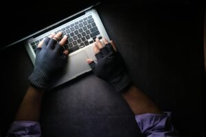 Immagine con visuale dall'alto, in stanza buia, di mani illuminate, ricoperte da guanti con la punta delle dita libere, che digitano sulla tastiera di un pc portatile (MacBook)