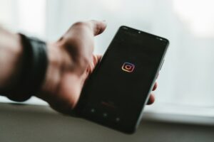 Immagine di una mano che regge un cellulare che si sta aprendo sull'app di Instagram, infatti la schermata è nera con solo il logo centrale.