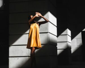 Immagine che rappresenta una donna con un vestito estivo giallo che si sopra il viso dal sole. Immagine con forti contrasti luce e ombra.